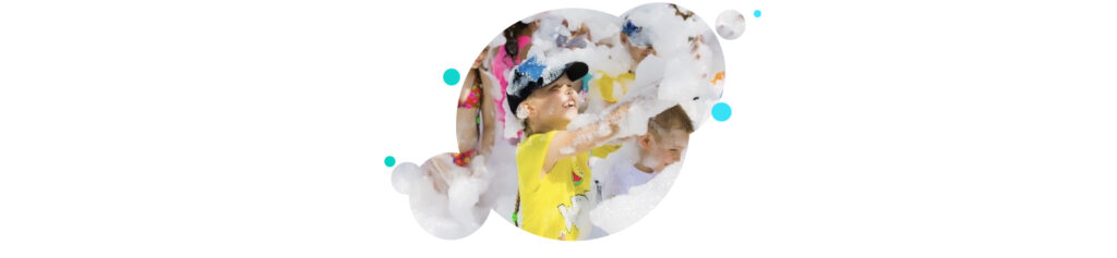 A child holds out their hands to catch foam. They're covered in foam and wearing a tshirt and shorts.